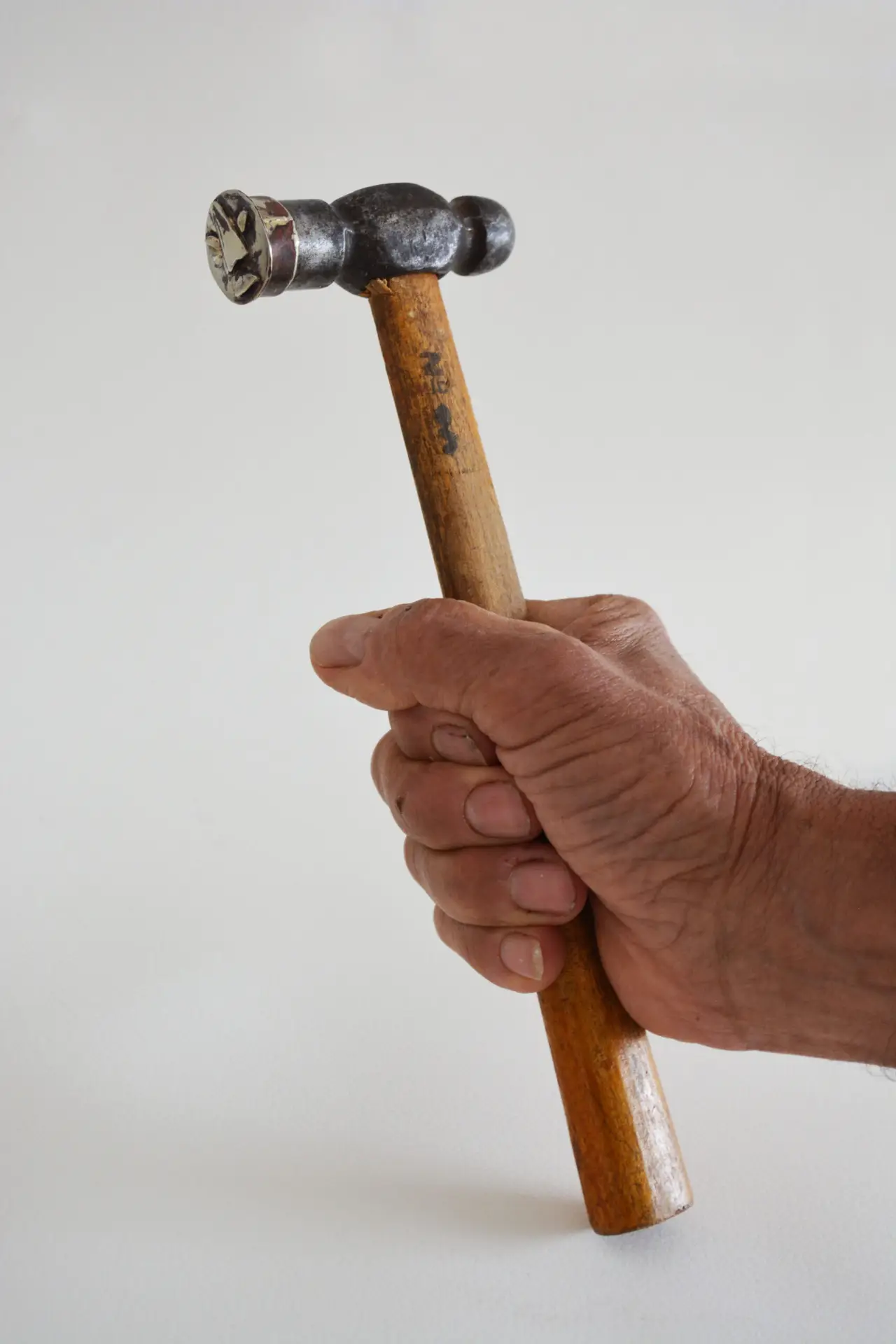 FACIA DA TOLA (2024): hand holding a hammer with a silver relief on its striking face, white background