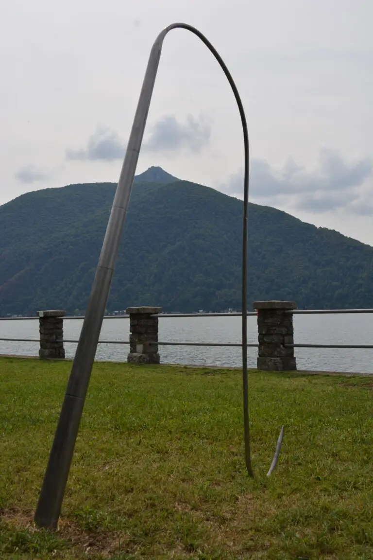 BIAS (2022): bent metal pole arching over a lawn by a lake, a mountain behind