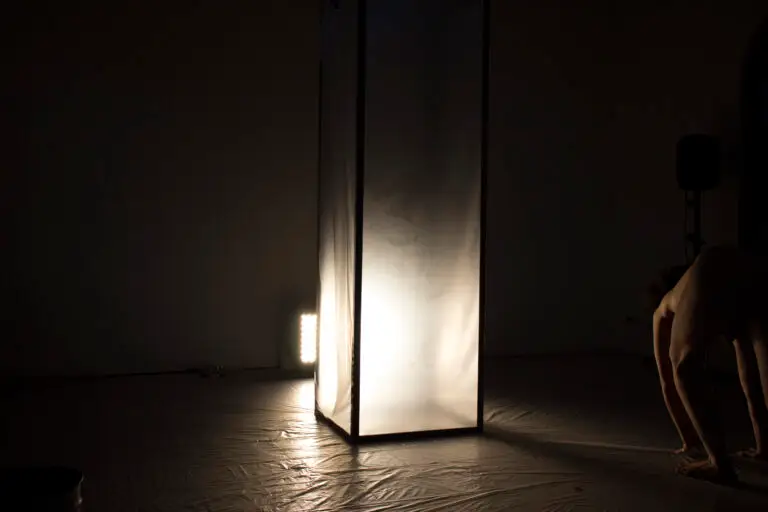 THICKEN UP (2022): tall translucent box lit from inside in a dark room, a crouching performer at the edge