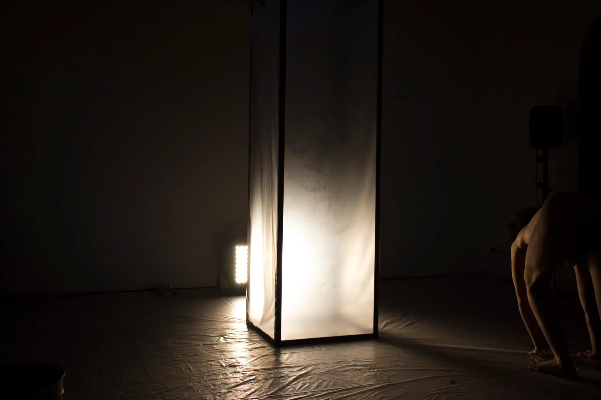 THICKEN UP (2022): tall translucent box lit from inside in a dark room, a crouching performer at the edge