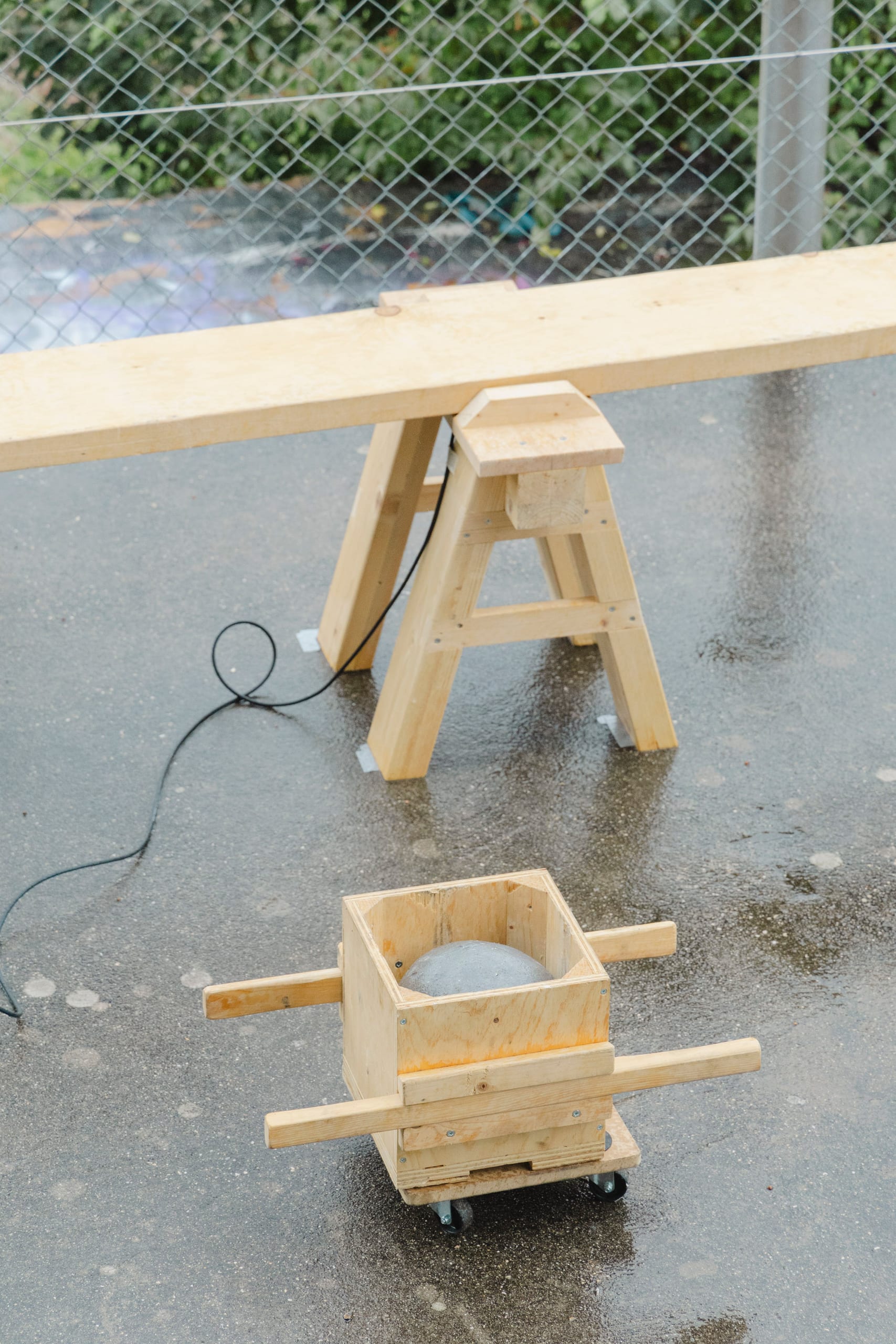 Il gioco è bello quando dura poco (2023): plank on a wooden trestle and a lead ball in a wooden carrier on wet asphalt